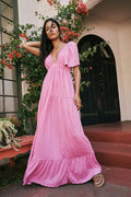 Woman in a pink dress standing outdoors with plants and flowers around.