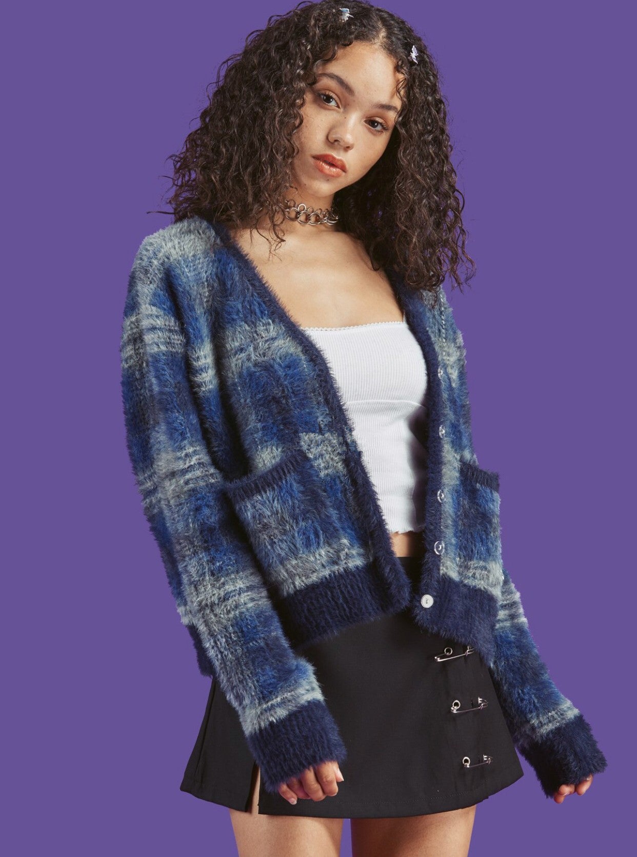 Woman wearing a blue plaid cardigan over a white top and black skirt on a purple background